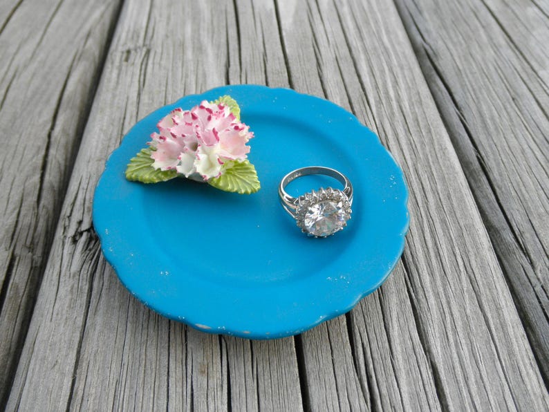 May include: A blue ceramic ring dish with a white and pink flower and a silver ring with a large clear stone.