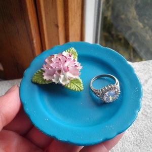 May include: A small blue ceramic dish with a white and pink flower and a silver ring with a large clear stone.