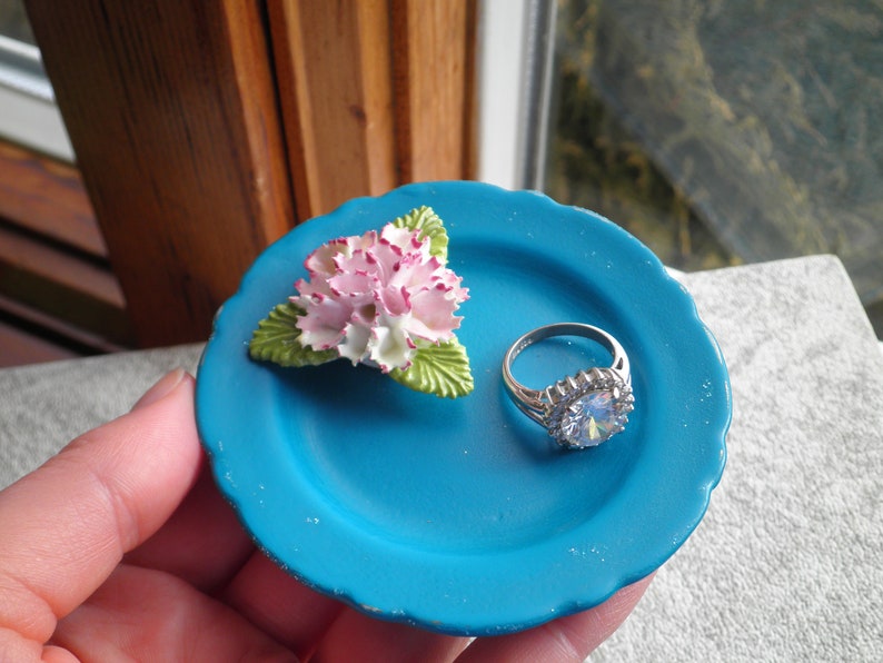 May include: A small blue ceramic dish with a white and pink flower and a silver ring with a large clear stone.