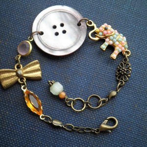 May include: A gold chain bracelet with a pink and blue elephant charm, a brown button, a gold bow, and a clear and orange crystal.