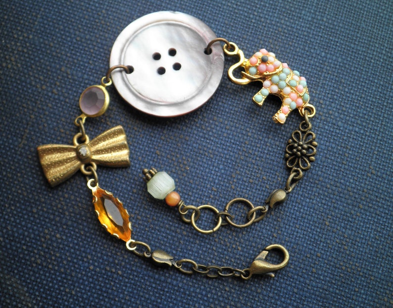 May include: A gold chain bracelet with a pink and blue elephant charm, a brown button, a gold bow, and a clear and orange crystal.