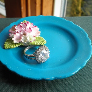 May include: A turquoise blue ceramic ring dish with a white and pink porcelain flower and a silver ring with a large clear stone.