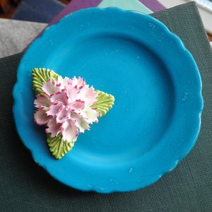 May include: A small, blue ceramic plate with a white and green ceramic flower on top. The plate has a scalloped edge.