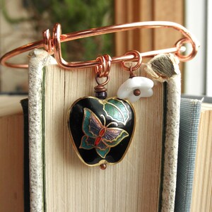 May include: A copper wire bracelet with a black heart charm featuring a butterfly design. The charm is decorated with green, blue, and red enamel. The bracelet also has a white flower charm.
