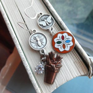 May include: Silver-toned dangle earrings with nature-inspired charms. One earring features a butterfly, the other a bee. Additional charms include a clear crystal and a small copper-colored plant pot. A colorful floral charm is also present.