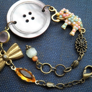 May include: A bracelet with a large, iridescent button, a small pink and blue elephant charm, a gold bow, and a variety of other beads.