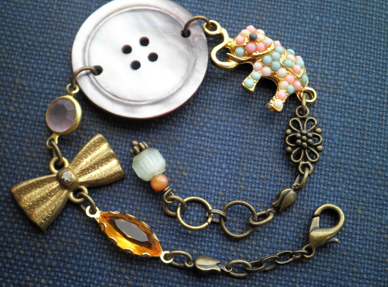 May include: A bracelet with a large, iridescent button, a small pink and blue elephant charm, a gold bow, and a variety of other beads.