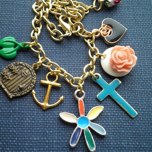 May include: A gold-toned charm bracelet with various pendants. Charms include a green leaf, a door, an anchor, a flower, a cross, a heart with a rose, and a pink rose. The bracelet is on a blue textured surface.