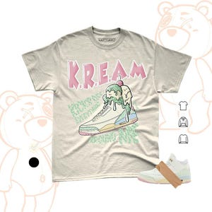 May include: A light beige t-shirt with the word "KREAM" in pink block letters. The shirt features a graphic of a sneaker with ice cream dripping over it. The brand name "NASTYJAMZ" is printed on the collar.