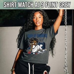 May include: Dark gray t-shirt with the text "SELF MADE" and a cartoon graphic. The graphic features a woman with a handbag, lipstick, and sneakers. The shirt is paired with black shorts.