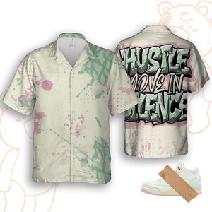 Könnte beinhalten: Ein kurzärmeliges Hemd mit cremefarbenem Grund, das rosa und grüne Farbspritzer aufweist. Auf der Rückseite des Hemdes steht der Text "Hustle Move in Silence" im Graffiti-Stil. Ein Paar weiße und rosa Turnschuhe ist ebenfalls abgebildet.