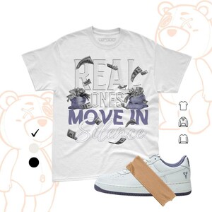 May include: White t-shirt with the text "REAL ONES MOVE IN SILENCE" in a stylized font, featuring money bag graphics and falling dollar bills. A pair of white and blue sneakers with a brown paper sleeve are also in the image.
