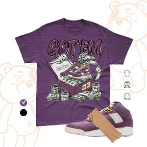 Könnte beinhalten: Ein violettes Kurzarm-T-Shirt mit einer Grafik, die einen Schuhkarton voller Geldscheine und ein Paar violette Sneaker zeigt. Der Text GOT'EM steht über dem Karton und die Zahl 23 befindet sich an der Seite. Ein passender violetter Sneaker ist darunter abgebildet.