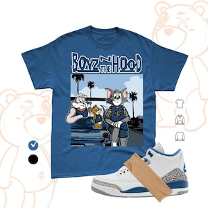May include: A blue short-sleeve t-shirt featuring a graphic print of cartoon characters Tom and Jerry with the text "BOYZ N THE HOOD" above them. The design includes palm trees and a car in the background. A white and blue sneaker is also shown.