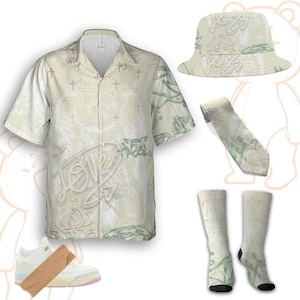 Könnte beinhalten: Ein koordiniertes Mode-Set mit einem kurzärmeligen Hemd, einem Fischerhut, einer Krawatte, Socken und Turnschuhen. Die Artikel haben eine cremefarbene Basis mit einem sich wiederholenden Kreuzmuster und Graffiti-Text und grünen Akzenten. Die Turnschuhe sind weiß mit rosa und hellblauen Akzenten.
