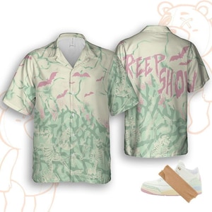 Könnte beinhalten: Ein kurzärmeliges Hemd mit hellgrünem und cremefarbenem Hintergrund, mit rosa Fledermäusen und Skelettfiguren. Auf der Rückseite des Hemdes steht in Rosa das Wort "CREEP SHOW". Ein Paar weiße und rosa Turnschuhe ist ebenfalls abgebildet.