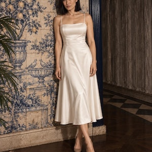 May include: A midi-length ivory satin dress featuring a fitted bodice, a straight neckline, and thin spaghetti straps. The skirt has a flared silhouette that falls below the knees, paired with neutral-toned pointed-toe heels.