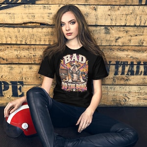 以下が含まれることがあります： サングラスとヘッドフォンを着用したブルドッグがレコードプレーヤーの横に座っているグラフィックが描かれた黒の半袖Tシャツ。BAD TO THE BONEという文字がかすれたフォントで書かれており、カラフルな星、音符、レコードが周囲に配置されています。