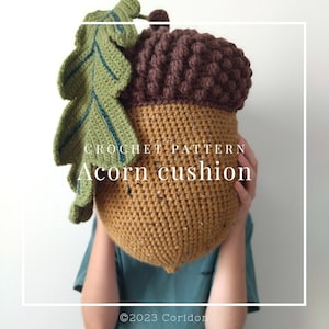 Op de afbeelding: Haakpatroon voor een eikel kussen. Het kussen is bruin en beige met een groen blad. De tekst "CROCHET PATTERN Acorn cushion" is zichtbaar op het kussen.