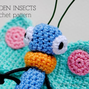 Op de afbeelding: Een gehaakte vlinder met blauwe vleugels, een oranje lichaam en roze en witte accenten. De vlinder heeft grote witte ogen met zwarte pupillen. De tekst "GARDEN INSECTS crochet pattern" is zichtbaar boven de vlinder.
