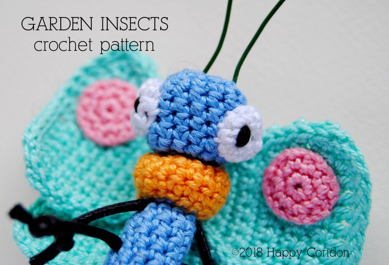 Op de afbeelding: Een gehaakte vlinder met blauwe vleugels, een oranje lichaam en roze en witte accenten. De vlinder heeft grote witte ogen met zwarte pupillen. De tekst "GARDEN INSECTS crochet pattern" is zichtbaar boven de vlinder.