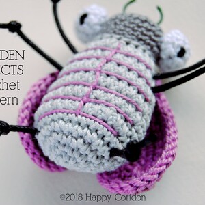 Op de afbeelding: Een gehaakt grijs en roze insect met zwarte poten en witte ogen. Het insect zit op een roze gehaakte basis. De tekst "GARDEN INSECTS crochet pattern" is zichtbaar in de afbeelding.