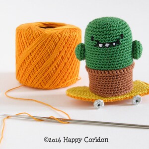 May include: A green crocheted cactus wearing a toothy smile, riding a yellow skateboard with white wheels. A ball of orange yarn and a crochet hook are in the background.