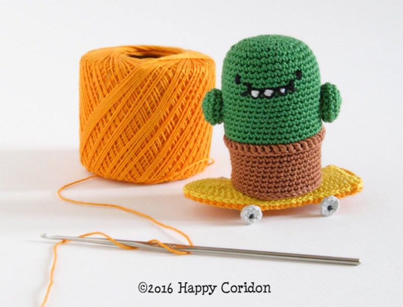 May include: A green crocheted cactus wearing a toothy smile, riding a yellow skateboard with white wheels. A ball of orange yarn and a crochet hook are in the background.