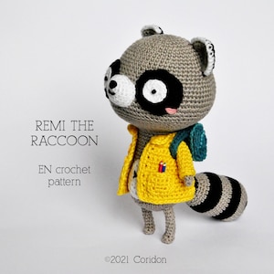 Puede incluir: Patrón de crochet para un mapache gris que lleva una chaqueta amarilla y una mochila turquesa. El patrón se llama "Remi el Mapache" y está escrito en inglés.