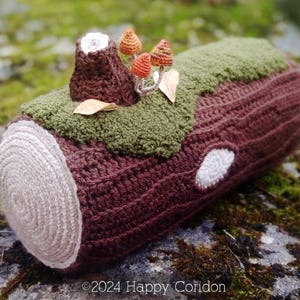May include: A crocheted log with a brown bark texture and a beige cross-section. The log is covered in green moss and has a small brown stump with orange and red mushrooms on top. The log is sitting on a mossy rock.
