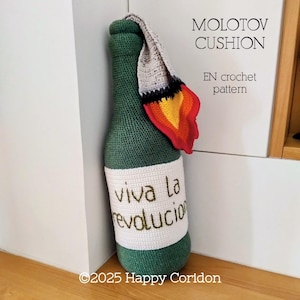 May include: A green crocheted bottle-shaped cushion with a white label that reads "Viva la revolucion" in green thread. A crocheted flame in red, yellow, and orange is attached to the top. The text "MOLOTOV CUSHION" and "EN crochet pattern" are also visible.