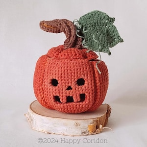 Pode incluir: Uma abóbora laranja de crochê com um rosto esculpido, um caule marrom e uma folha verde. A abóbora está em uma fatia de madeira. O texto "©2024 Happy Coridon" está na parte inferior.
