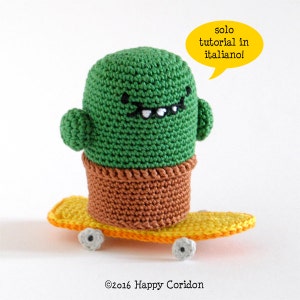 May include: A green crocheted cactus wearing a brown pot and riding a yellow skateboard with black wheels. The cactus has a smiling face with black eyes and a white mouth. The text "solo tutorial in italiano!" is in a yellow speech bubble above the cactus.