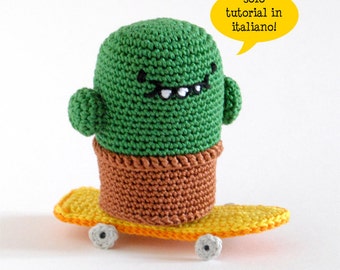 Può includere: Un cactus verde all'uncinetto che indossa un vaso marrone e che sta su uno skateboard giallo con ruote nere. Il cactus ha un viso sorridente con occhi neri e una bocca bianca. Il testo "solo tutorial in italiano!" è in un fumetto giallo sopra il cactus.