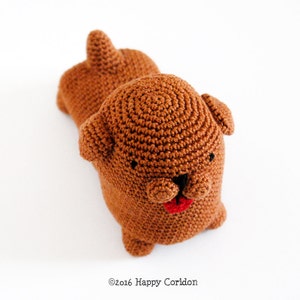 May include: A brown crocheted dog toy with black eyes and a red tongue. The dog is lying on its side with its head turned to the side.