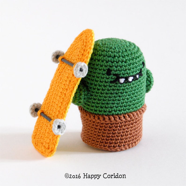 May include: A green crocheted cactus with a smiling face and a black mustache, holding a yellow skateboard with black wheels. The cactus is wearing a brown crocheted pot.