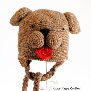 May include: Brown crocheted dog hat with a red tongue and black eyes. The hat has a long brown braided strap.