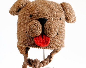 Può includere: Cappello da cane in crochet marrone con una lingua rossa e occhi neri. Il cappello ha un lungo cinturino intrecciato marrone.