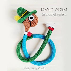 Puede incluir: Un juguete de gusano tejido a ganchillo con un sombrero verde, una pluma amarilla y un cuerpo azul y verde. El gusano tiene una cabeza marrón con un ojo grande y una pajarita roja. El texto "LOWLY WORM En crochet pattern" es visible.