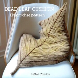 May include: A leaf-shaped cushion, crocheted in tan and brown yarn, displayed on a white chair. The cushion's design includes detailed stitching to resemble leaf veins. The image text reads "DEAD LEAF CUSHION EN crochet pattern" and "©2024 Coridon".
