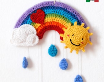 Op de afbeelding: Een gehaakte regenboogmobiel met een witte wolk, een gele zon met een smiley en blauwe regendruppels die eraan hangen. De regenboog is gemaakt van verschillende gekleurde garens, en de zon heeft een vrolijk gezicht.