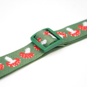 May include: Green strap with a repeating pattern of red and white mushrooms. A green plastic buckle is attached to the strap. The strap is likely for a pet collar or accessory.