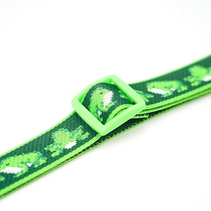 May include: Green and white frog-patterned strap with a lime green square buckle. The strap is dark green with repeating pixelated frog designs. The buckle is a solid, bright green color. The strap is likely for a bag or accessory.