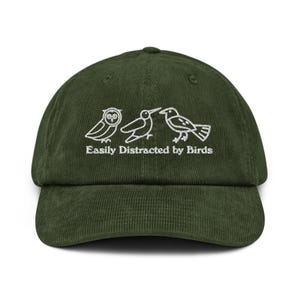 Puede incluir: Gorra de béisbol de pana verde oscuro con visera curva. La gorra presenta un diseño bordado blanco de tres pájaros y el texto "Easily Distracted by Birds". Un accesorio casual.