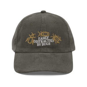 Puede incluir: Gorra de béisbol de pana verde oliva con el texto bordado "EASILY DISTRACTED BY BUGS" e ilustraciones de insectos en hilo dorado. La gorra tiene una visera curva y una correa ajustable.
