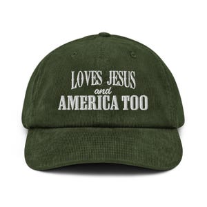 Pode incluir: Boné de beisebol de veludo cotelê verde escuro com o texto bordado em branco «LOVES JESUS and AMERICA TOO». O boné tem uma aba curva e uma coroa arredondada.