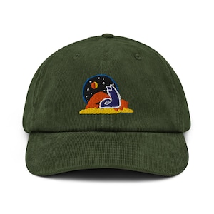 Op de afbeelding: Groene corduroy baseballpet met een geborduurd ontwerp van een huilende wolf onder een sterrenhemel. De wolf is blauw, de lucht is donkerblauw en het landschap is oranje en geel.