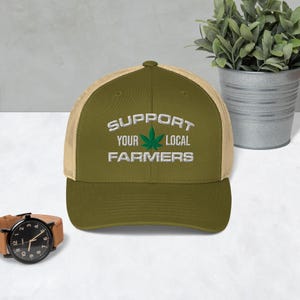 May include: Olive green trucker hat with the text "SUPPORT YOUR LOCAL FARMERS" in white, with a green cannabis leaf graphic. The hat has a tan mesh back and a curved brim. A wristwatch with a brown leather band is in the foreground.