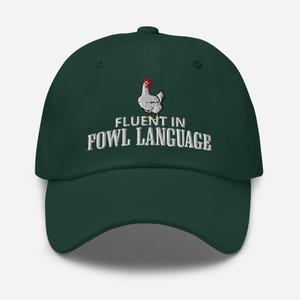 Può includere: Un cappello da baseball verde scuro con ricamo bianco che recita "Fluent in Fowl Language". Un pollo bianco con cresta rossa è ricamato sopra il testo.