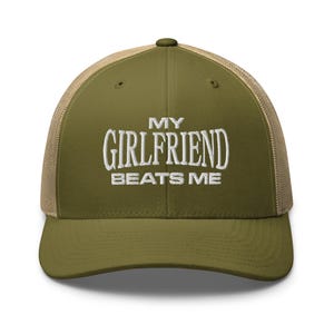 May include: A green and brown trucker hat with the text "My Girlfriend Beats Me" embroidered in white.